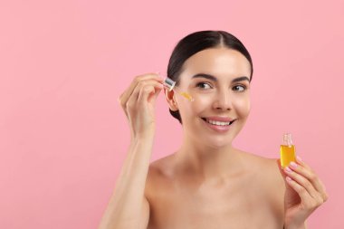 Güzel genç bir kadın pembe arka planda yüzüne serum uyguluyor. Metin için boşluk