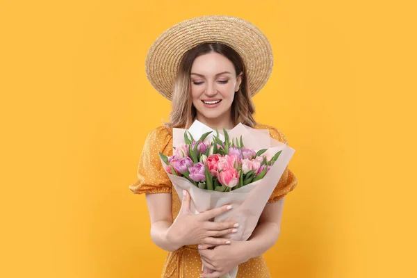 黄色の背景に美しいチューリップの花束を保持するわら帽子で幸せな若い女性 — ストック写真