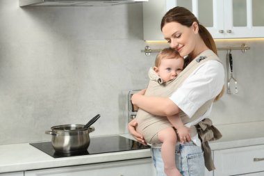 Anne mutfakta çocuğunu askıda (bebek taşıyıcısı) tutuyor.