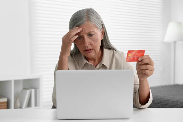 Upset woman with credit card near laptop at table indoors. Be careful - fraud