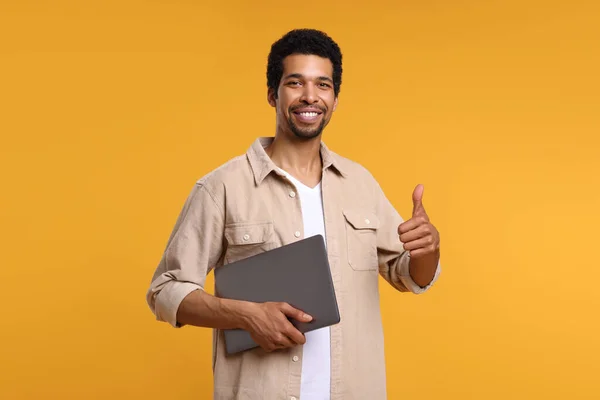 オレンジ色の背景に親指を示すラップトップを持つ幸せな男 — ストック写真