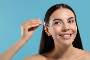 Güzel genç bir kadın açık mavi arka planda yüzüne serum sürüyor.