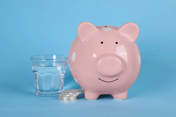 Water scarcity concept. Piggy bank, coins and glass of drink on light blue background