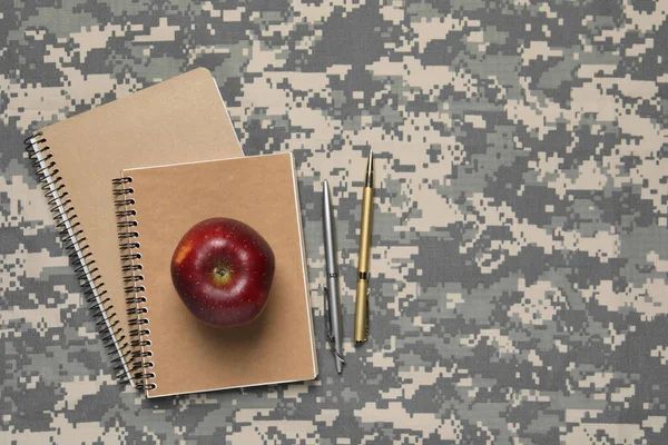 Notebooks, pens and apple on camouflage background, flat lay with space for text. Military education