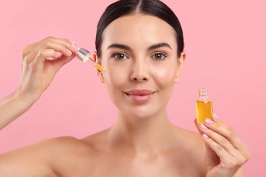 Güzel genç bir kadın pembe arka planda yüzüne serum sürüyor.