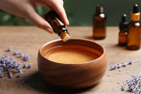 Woman dripping essential oil from bottle into bowl near lavender at wooden table, closeup