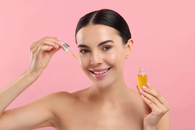 Güzel genç bir kadın pembe arka planda yüzüne serum sürüyor.