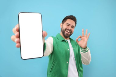 Genç adam elinde akıllı telefon ve açık mavi arka planda iyi bir jest gösteriyor.