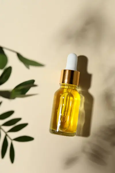 Bottle of cosmetic oil and leaves on beige background, top view