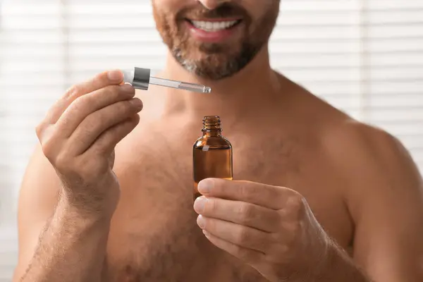 Smiling man with cosmetic serum indoors, selective focus