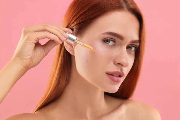 stock image Beautiful young woman applying cosmetic serum onto her face on pink background