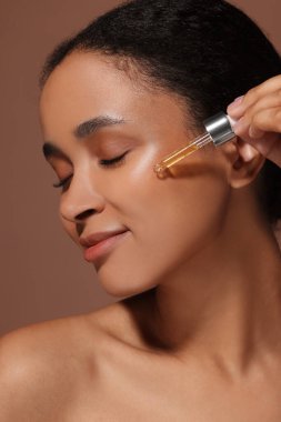Güzel bir kadın kahverengi arka planda yüzüne serum sürüyor.