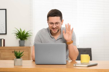 Evdeki ahşap masada bilgisayarla görüntülü konuşma yapan bir adam.