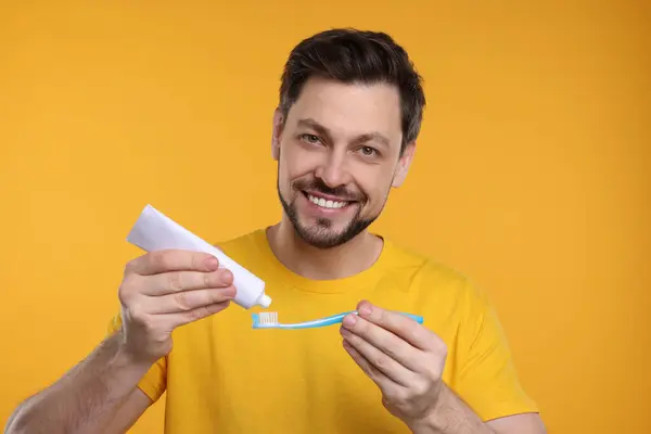 Happy man squeezing toothpaste from tube onto plastic toothbrush on yellow background