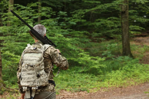 Man with hunting rifle wearing camouflage in forest, back view. Space for text