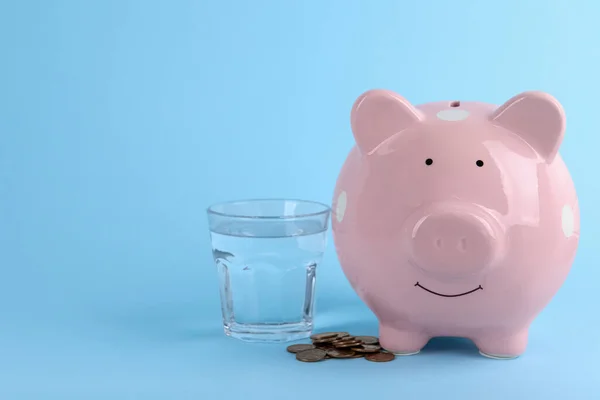 Water scarcity concept. Piggy bank, glass of drink and coins on light blue background, space for text