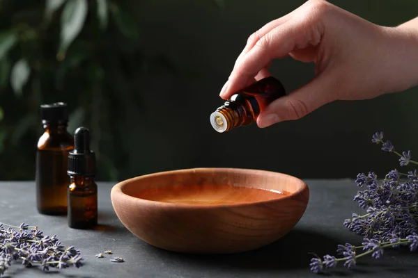 Woman dripping essential oil from bottle into bowl near lavender at grey table, closeup