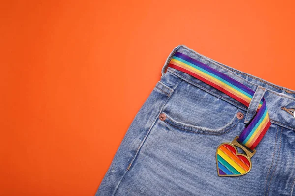 Jeans and rainbow ribbon with heart shaped pendant on orange background, top view and space for text. LGBT pride