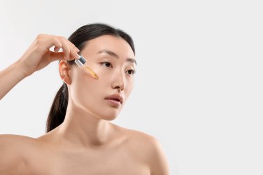 Güzel genç bir kadın yüzüne beyaz arka planda kozmetik serumu uyguluyor.