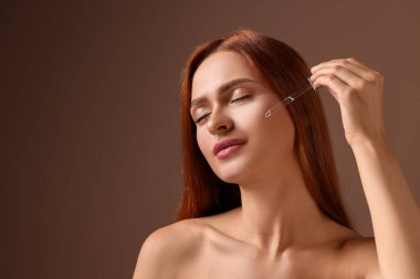Güzel genç bir kadın yüzüne kozmetik serumu sürüyor. Kahverengi arka planda, mesaj için yer var.