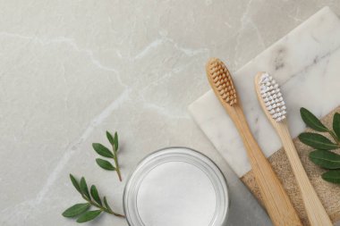 Bambu diş fırçaları, kabartma tozu dolu cam kase ve açık gri mermer masa üzerinde yeşil yapraklar. Metin için boşluk
