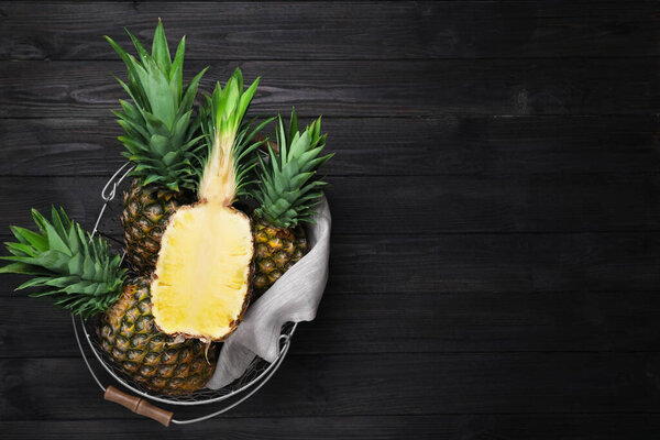Whole and cut ripe pineapples in metal basket on black wooden table, top view. Space for text
