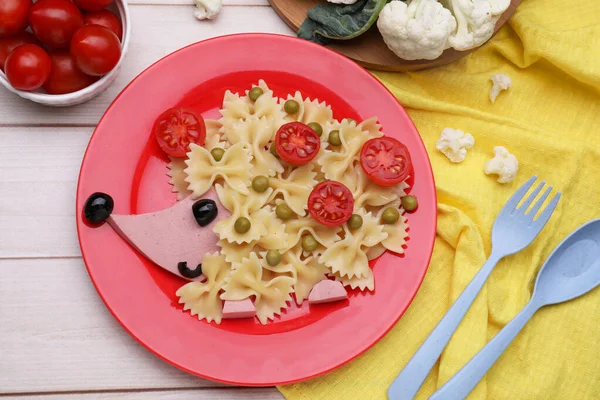 Creative serving for kids. Plate with cute hedgehog made of delicious pasta, sausages and tomatoes on white wooden table, flat lay