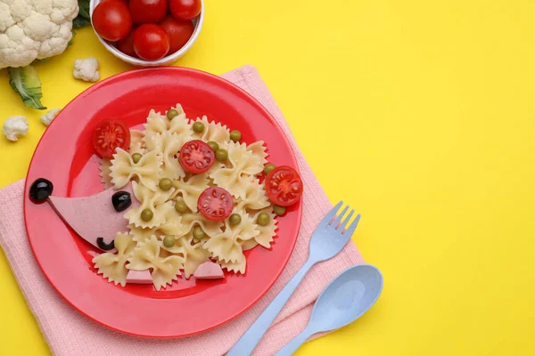 Creative serving for kids. Plate with cute hedgehog made of delicious pasta, sausages and tomatoes on yellow table, flat lay. Space for text