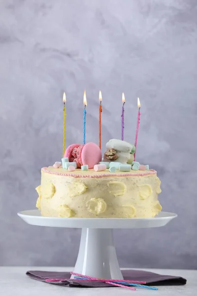 Delicious cake decorated with macarons, marshmallows and burning candles served on white table against grey background