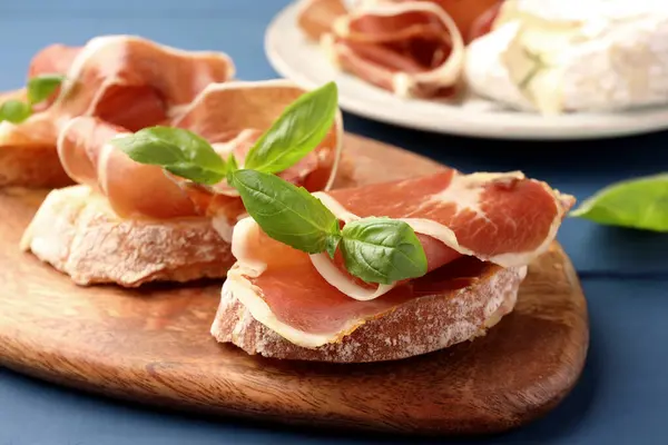 Tasty sandwiches with cured ham and basil leaves on blue wooden table, closeup