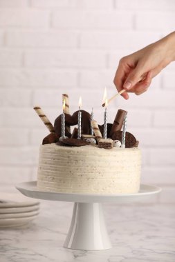 Beyaz mermer masadaki şekerlerle süslenmiş pastanın üzerinde mum yakan kadın.