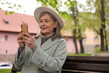 Güzel, son sınıf öğrencisi bir kadın bankta oturuyor ve açık havada akıllı telefon kullanıyor.