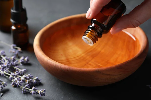 Woman dripping essential oil from bottle into bowl near lavender at grey table, closeup