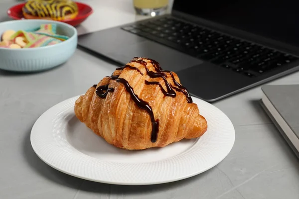 Bad eating habits at workplace. Delicious croissant and laptop on grey table, closeup