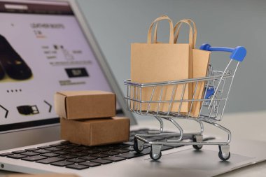 Mini alışveriş çantaları, el arabası ve kutular dizüstü bilgisayarda açık gri arka planda, yakın plan. Çevrimiçi mağaza