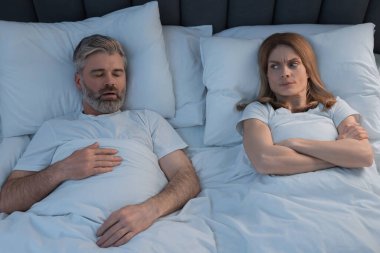 Evde yatakta horlayan kocasının yanında yatan sinirli kadın.