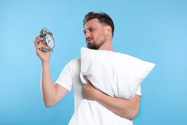 Unhappy man with pillow and alarm clock on light blue background. Insomnia problem