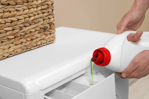 Man pouring fabric softener from bottle into washing machine indoors, closeup