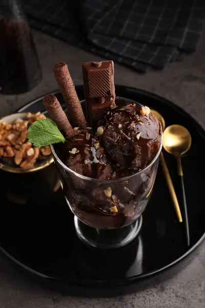 Tasty chocolate ice cream with sauce, nuts and wafer rolls in glass dessert bowl on dark grey table