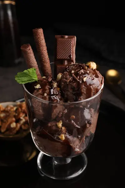 Tasty chocolate ice cream with sauce, nuts and wafer rolls in glass dessert bowl on table, closeup