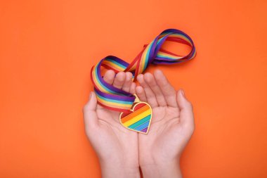 Turuncu arka planda kalp şeklinde kolyesi olan gökkuşağı kurdelesi tutan bir kadın. LGBT gururu