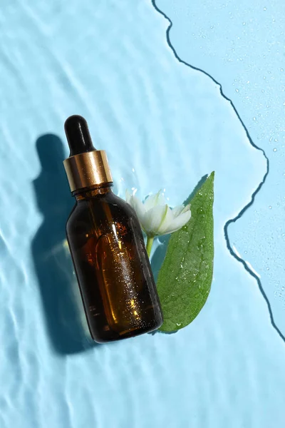 Bottle of cosmetic oil, green leaf and flower in water on light blue background, flat lay