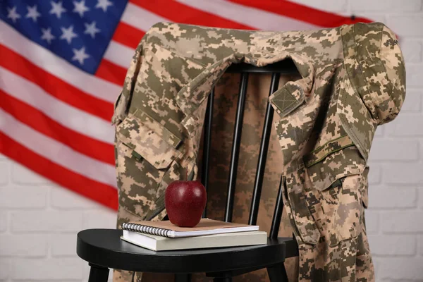 Soldier uniform, notebooks and apple on chair near flag of United States. Military education