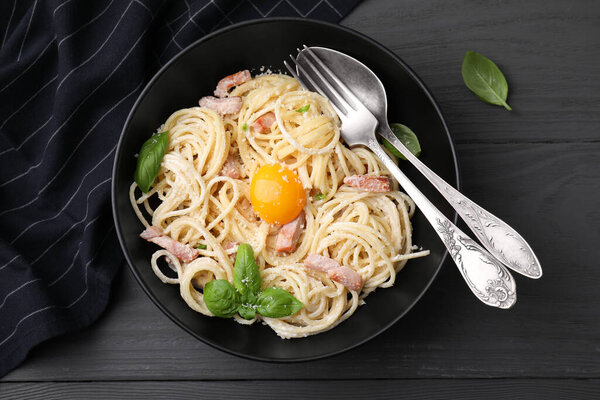 Bowl of delicious pasta Carbonara with egg yolk and cutlery on black wooden table, flat lay