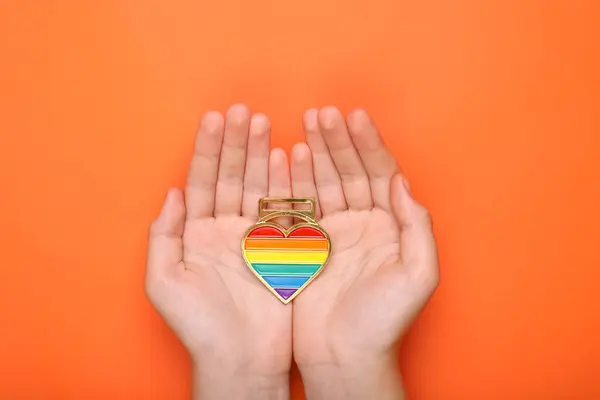 Woman holding rainbow heart shaped pendant on orange background, top view. LGBT pride