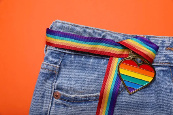 Jeans and rainbow ribbon with heart shaped pendant on orange background, top view. LGBT pride