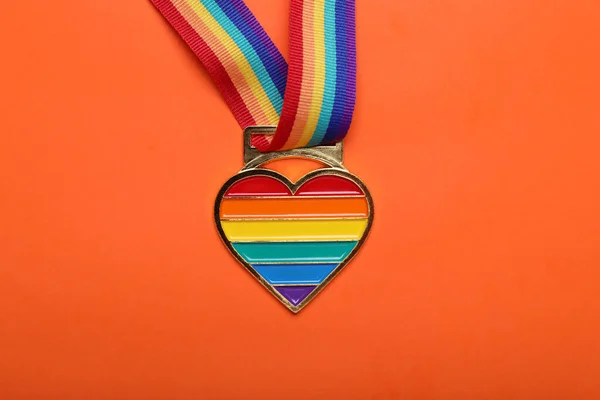 Rainbow ribbon with heart shaped pendant on orange background, top view. LGBT pride