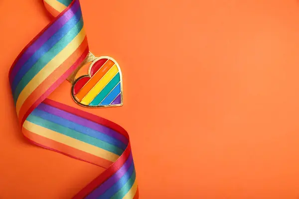 Rainbow ribbon with heart shaped pendant on orange background, top view and space for text. LGBT pride