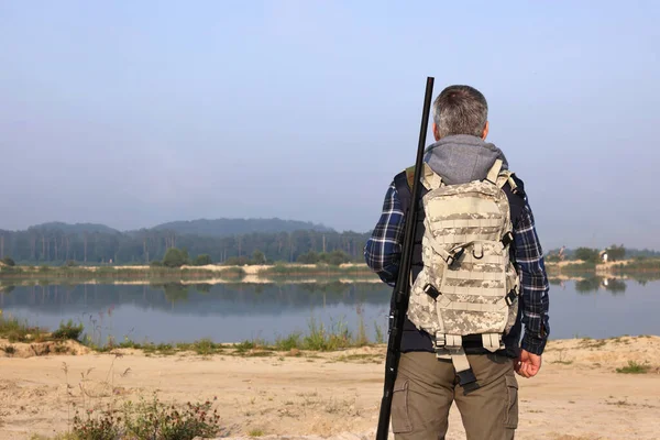 Man with hunting rifle and backpack near lake outdoors, back view. Space for text
