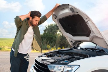 Kırık bir arabanın kaputunun altına bakan üzgün bir adam.